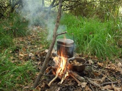 Dampfender Topf an einem eingegrabenen Ast hängt über einem Lagerfeuer. In der Glut steht ein weiterer Topf mit kochenden Kartoffeln.